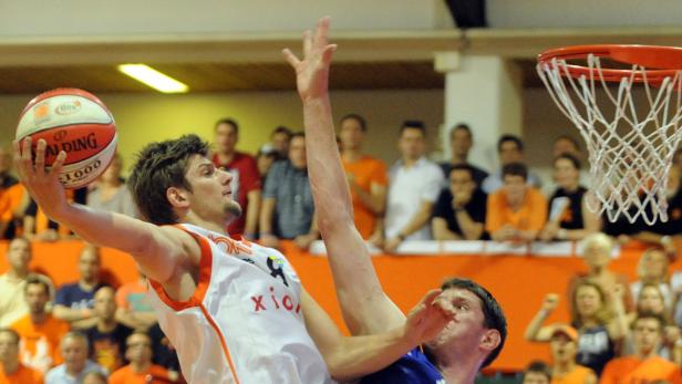 Ein Basketballspieler springt, um den Ball in den Korb zu werfen, während ein anderer Spieler ihn zu blocken versucht.