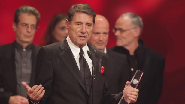 Ein Mann im Anzug hält eine Auszeichnung in der Hand und spricht vor Publikum.