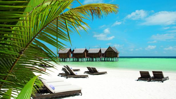 Strand mit Liegestühlen und Wasserbungalows unter blauem Himmel.