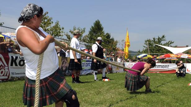 Frauen und Männer in Kilts beim Tauziehen auf einer Wiese.