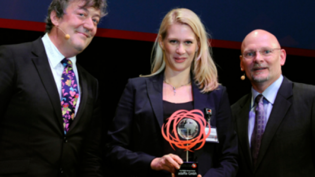 Eine blonde Frau hält eine Auszeichnung zwischen Stephen Fry und einem weiteren Mann.