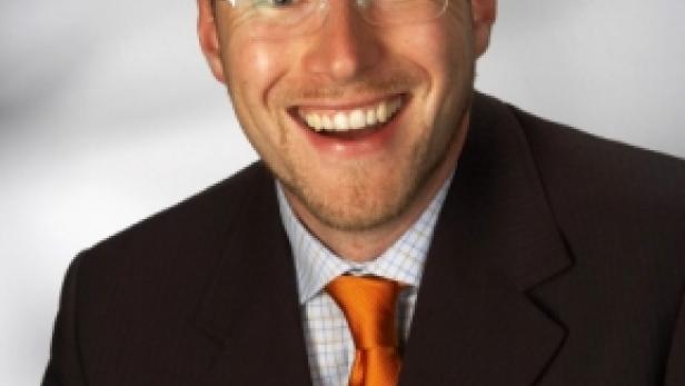 Ein lächelnder Mann mit Brille, Anzug und orangefarbener Krawatte vor einem grauen Hintergrund.