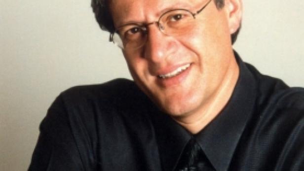 Ein lächelnder Mann mit Brille und dunklem Hemd posiert mit verschränkten Armen.
