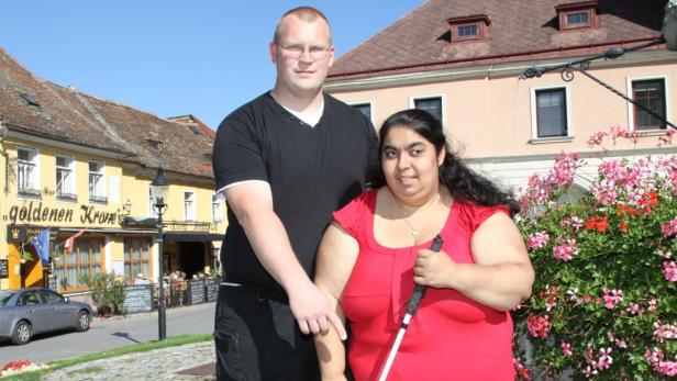 Ein Mann begleitet eine blinde Frau mit Blindenstock in einer Stadt.
