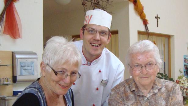 Ein Koch mit zwei älteren Damen in einem hellen Raum.