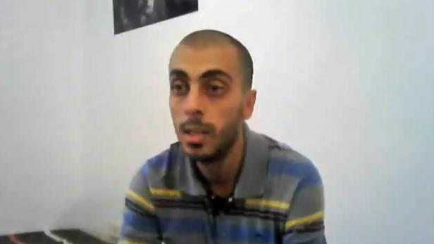 Ein Mann mit rasiertem Kopf und gestreiftem Hemd spricht vor einer weißen Wand.
