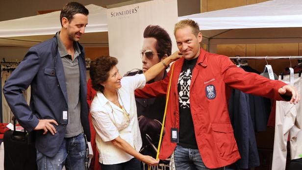 Eine Frau nimmt Maß an einem Mann, der eine rote Jacke mit dem österreichischen Wappen trägt.