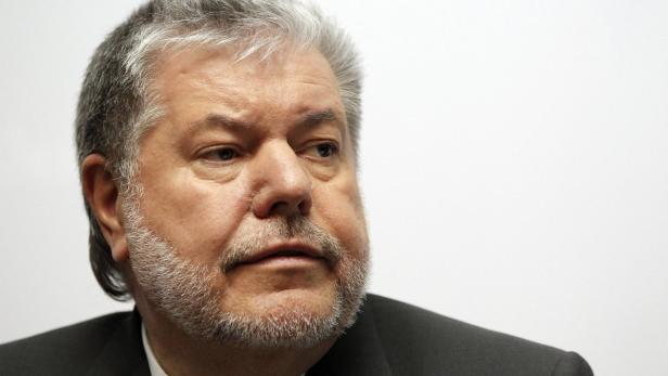 Ein Mann mit grauem Haar und Bart in einem dunklen Anzug blickt nach rechts.