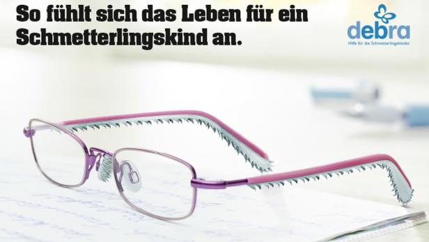 Eine Brille mit gezackten Bügeln liegt auf einem Blatt Papier; Werbung für die Organisation Debra.