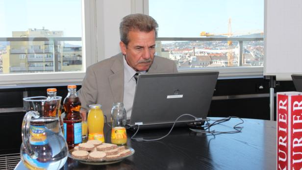 Ein Mann im Anzug sitzt an einem Tisch mit Laptop, Getränken und Keksen.