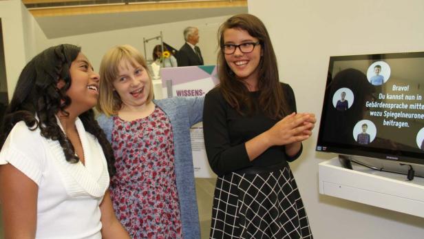 Drei junge Frauen stehen lächelnd vor einem Bildschirm mit Gebärdensprache-Informationen.