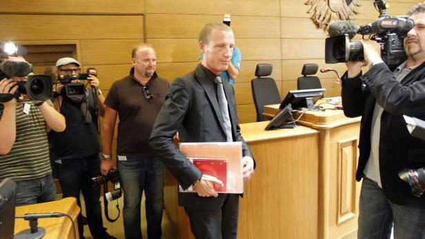 Ein Mann im Anzug verlässt einen Gerichtssaal, umringt von Kameraleuten.