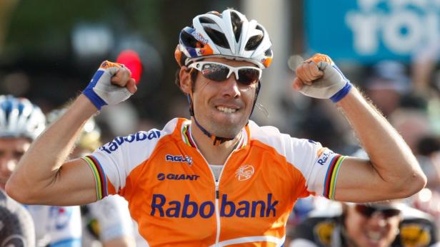 Ein Radrennfahrer im orangefarbenen Trikot jubelt mit erhobenen Fäusten.