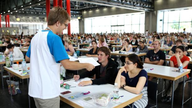 Ein Prüfer verteilt Dokumente an eine Gruppe von Studenten in einem großen Saal.