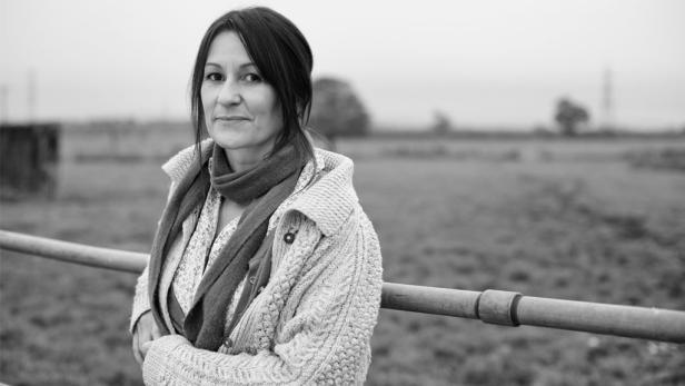 Eine Frau mit Schal und Strickjacke lehnt an einem Geländer auf dem Land.