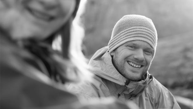 Ein lächelnder Mann mit Mütze und Jacke in Schwarzweiß.