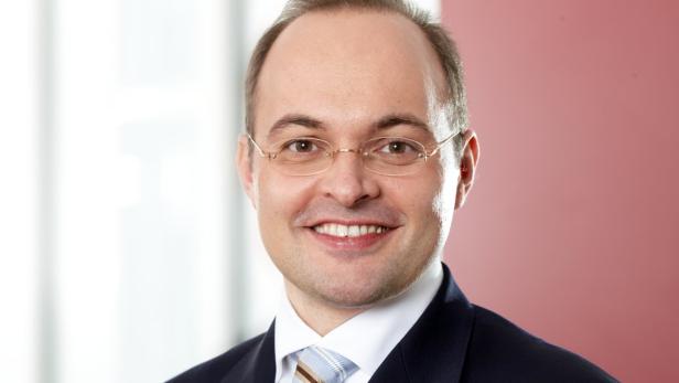 Ein lächelnder Mann mit Brille und Anzug vor einem roten Hintergrund.