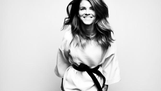 Eine lächelnde Frau mit wehendem Haar und einem Gürtel um eine helle Jacke.