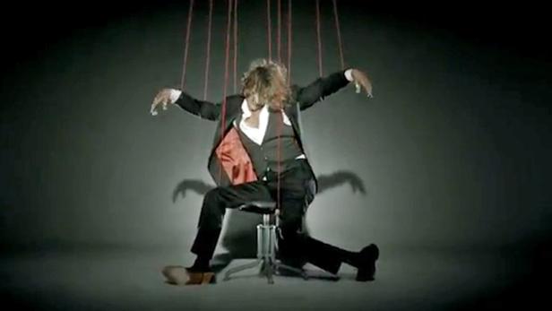 Ein Mann im Anzug sitzt wie eine Marionette an Fäden auf einem Stuhl.