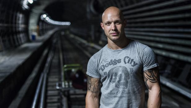 Ein Mann mit Tätowierungen steht in einem dunklen Tunnel mit Gleisen.