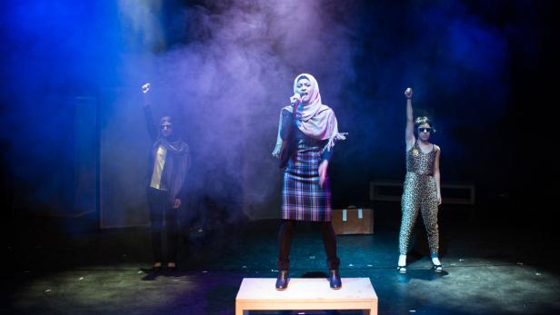 Eine Frau mit Hijab singt auf einer Bühne, flankiert von zwei Frauen mit erhobenen Fäusten.