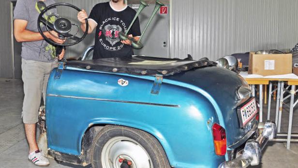 Zwei Männer präsentieren ein blaues Auto-Heck mit Lenkrad und Hupen.