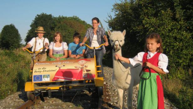Eine Gruppe von Kindern und eine Frau stehen mit einer Draisine und einem Alpaka auf einem Bahngleis.