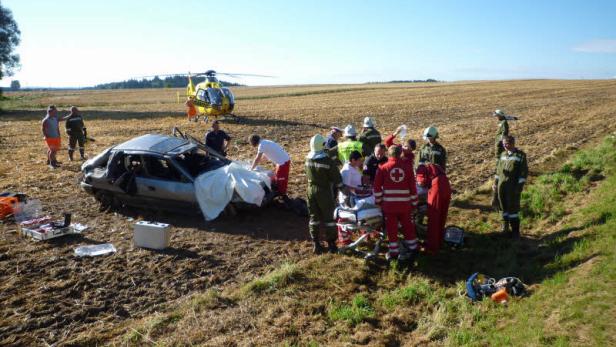 Nach einem Autounfall wird eine Person von Rettungskräften versorgt, während ein Hubschrauber bereitsteht.