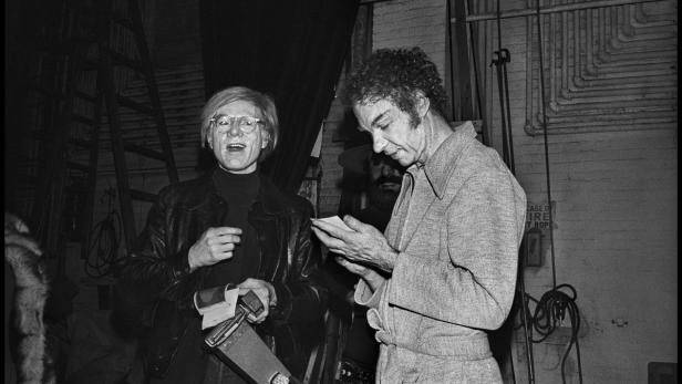 Andy Warhol und ein Mann mit lockigem Haar stehen zusammen in einem Raum.