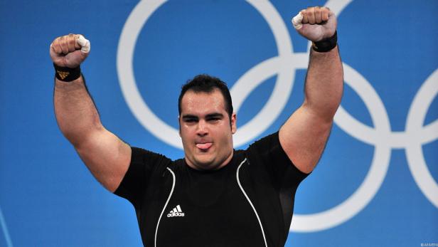 Ein Gewichtheber jubelt vor dem Hintergrund der Olympischen Ringe.
