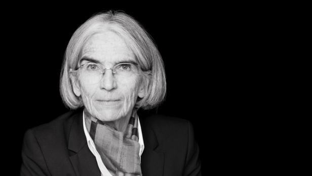 Schwarzweiß-Porträt einer älteren Frau mit Brille, Blazer und Schal vor schwarzem Hintergrund.