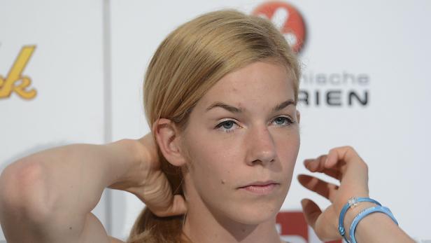 Eine blonde Frau mit blauen Augen fasst sich an den Hinterkopf.