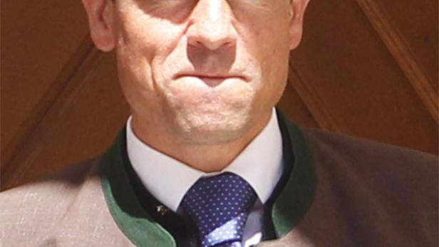 Ein Mann in Tracht mit Krawatte vor einer Holzwand.