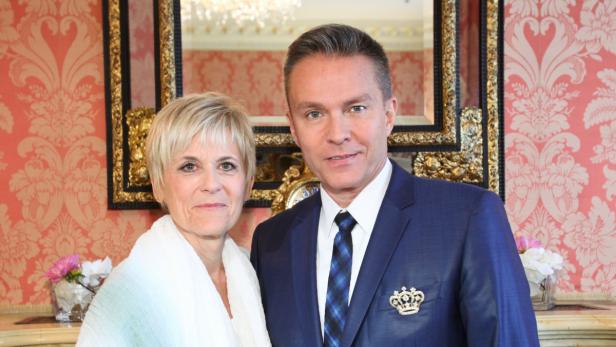 Ein Mann im Anzug mit einer Brosche und eine Frau stehen vor einer Wand mit Blumenmuster.