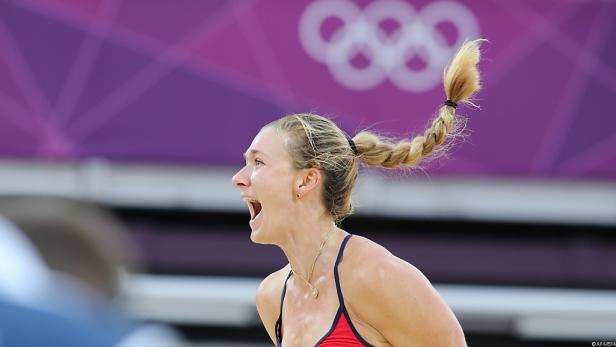 Eine Beachvolleyballspielerin jubelt mit offenem Mund vor dem Hintergrund der olympischen Ringe.