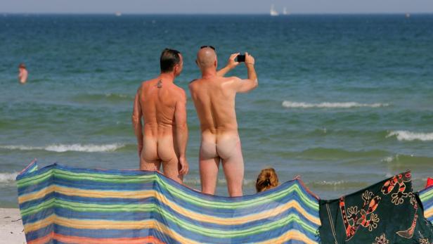 Zwei Männer stehen hüllenlos am Strand, einer fotografiert mit seinem Handy das Meer.