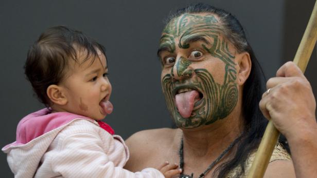 Ein Mann mit traditioneller Maori-Gesichtsbemalung hält ein Baby, beide strecken die Zunge heraus.