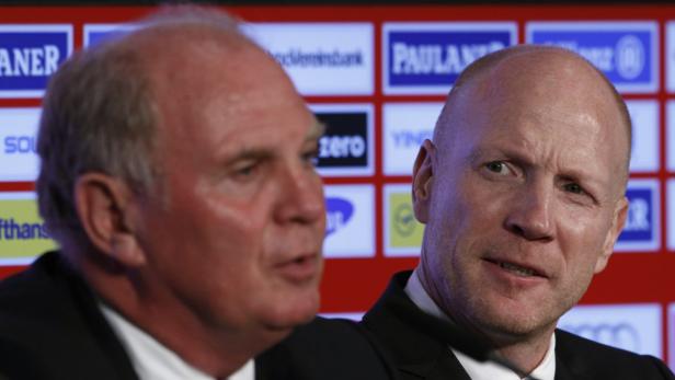 Zwei Männer in Anzügen stehen vor einer Wand mit Sponsorenlogos.