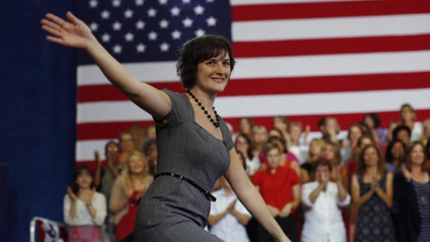Eine Frau in einem grauen Kleid winkt vor einer jubelnden Menge und der US-amerikanischen Flagge.