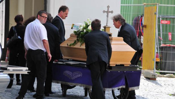 Mehrere Personen schieben einen Sarg auf einem Wagen durch die Straße.