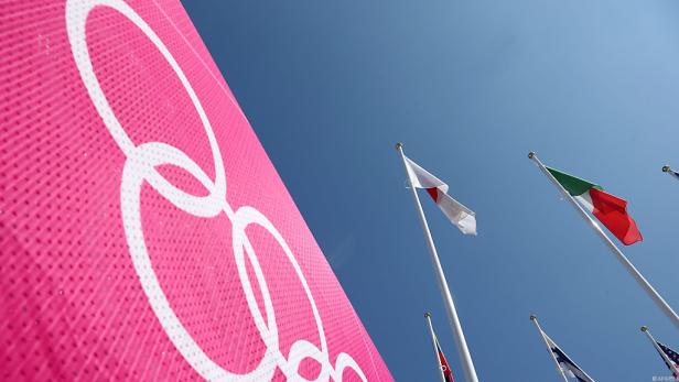 Olympische Ringe auf einem Banner, im Hintergrund wehen verschiedene Nationalflaggen.