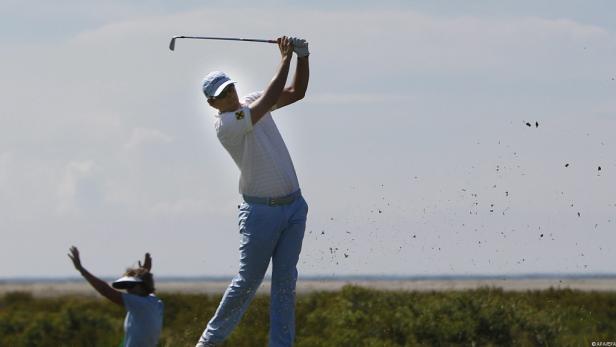 Ein Golfer schlägt den Ball, während eine Zuschauerin im Hintergrund steht.