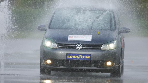 Ein grauer VW wird mit Wasser bespritzt, während er auf einer nassen Straße fährt.
