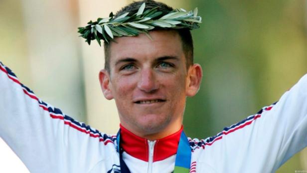 Ein Mann mit Lorbeerkranz und olympischer Medaille jubelt.