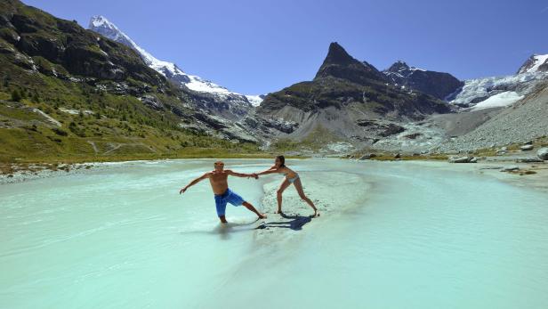 Ein Paar watet in einem türkisfarbenen Gletschersee vor einer Bergkulisse.