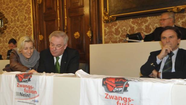 Mehrere Personen sitzen an einem Tisch mit einem Banner gegen eine „Zwangsfusion“.