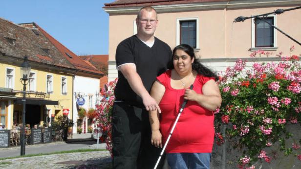 Eine blinde Frau mit Blindenstock steht mit einem Mann vor einem Gebäude mit Blumen.