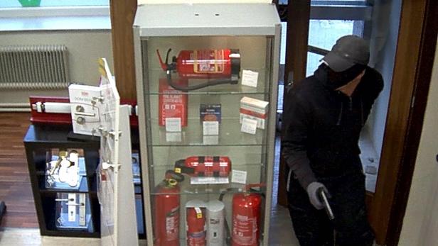 Eine vermummte Person steht neben einem Schaufenster mit Feuerlöschern.