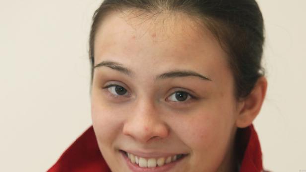 Eine lächelnde junge Frau mit dunklen Haaren und einem roten Kragen vor einem hellen Hintergrund.