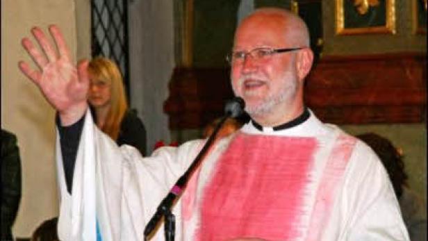 Ein Priester mit Brille und weiß-rotem Gewand spricht in ein Mikrofon.